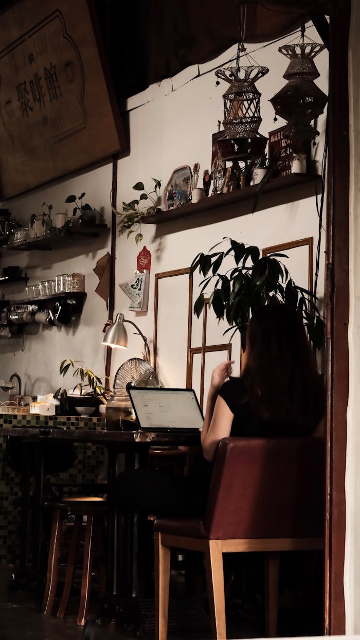 about-01 Woman working with a laptop in a cozy cafe surrounded by plants and warm decor.